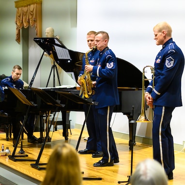 An Air Force alto saxophonist is seen flanked by two other members of the music ensemble as he performs a solo with a pianist who is seen in the far left of the picture. Each are wearing dark blue Air Force uniforms, and they are standing on a light colored wood stage.