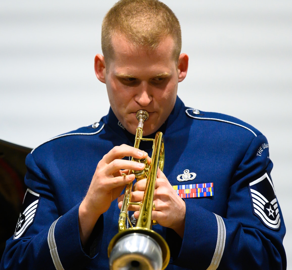 An Air Force trumpeter performs a solo using with a silver mute. He is wearing dark blue Air Force uniform against a white wall.