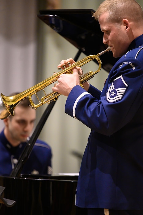 An Air Force trumpeter performs a solo and is pictures on the right, with a pianist who is seen in the far left of the picture. Each are wearing dark blue Air Force uniforms.