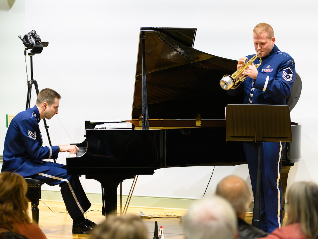 An Air Force trumpeter performs a solo and is pictures on the right, with a pianist who is seen in the far left of the picture. Each are wearing dark blue Air Force uniforms.