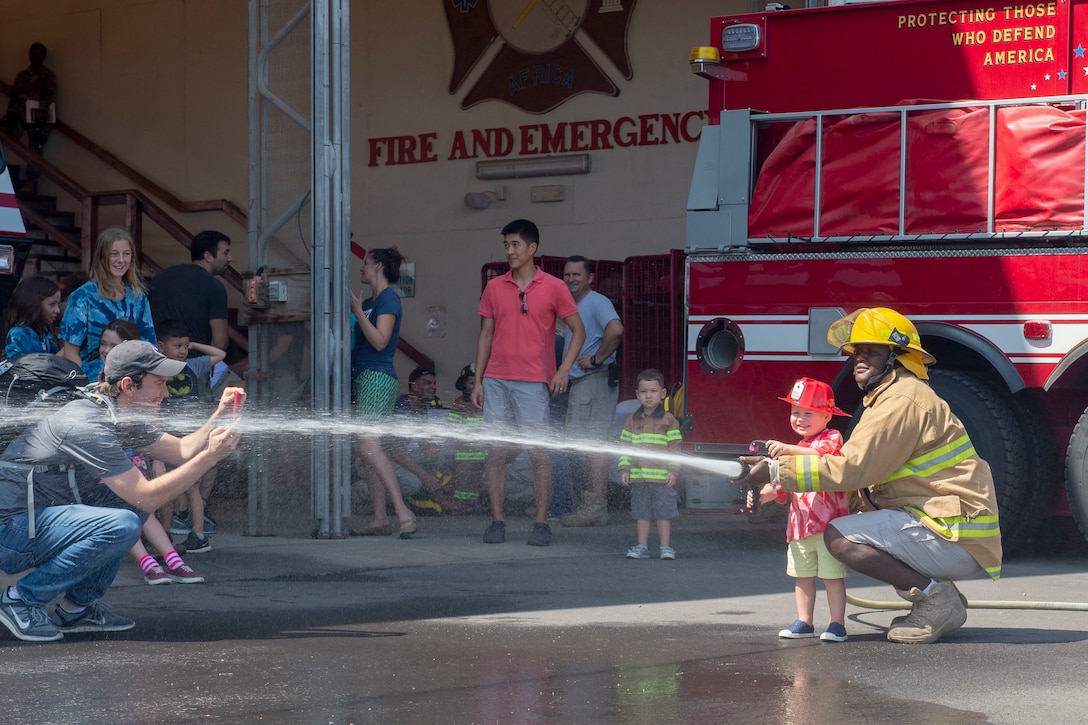 A firefighter helps a kid man a hose as a crowd of adults and kids watch.