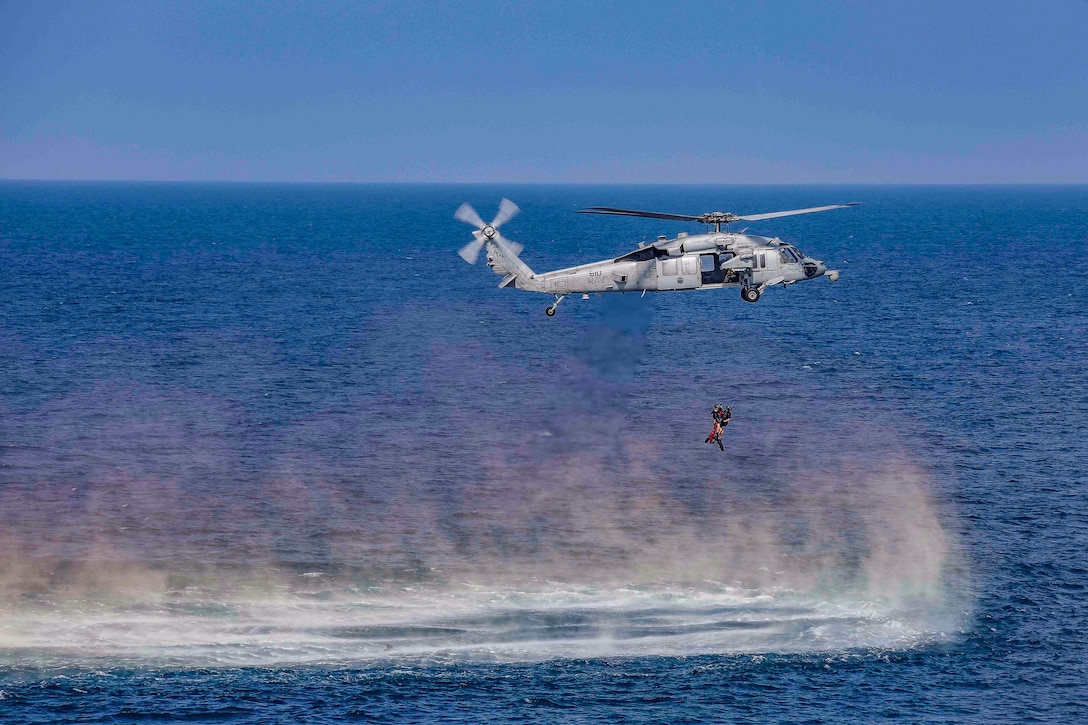 A low hovering helicopter kicks up sea spray from deep blue waters while hoisting service members on a line.