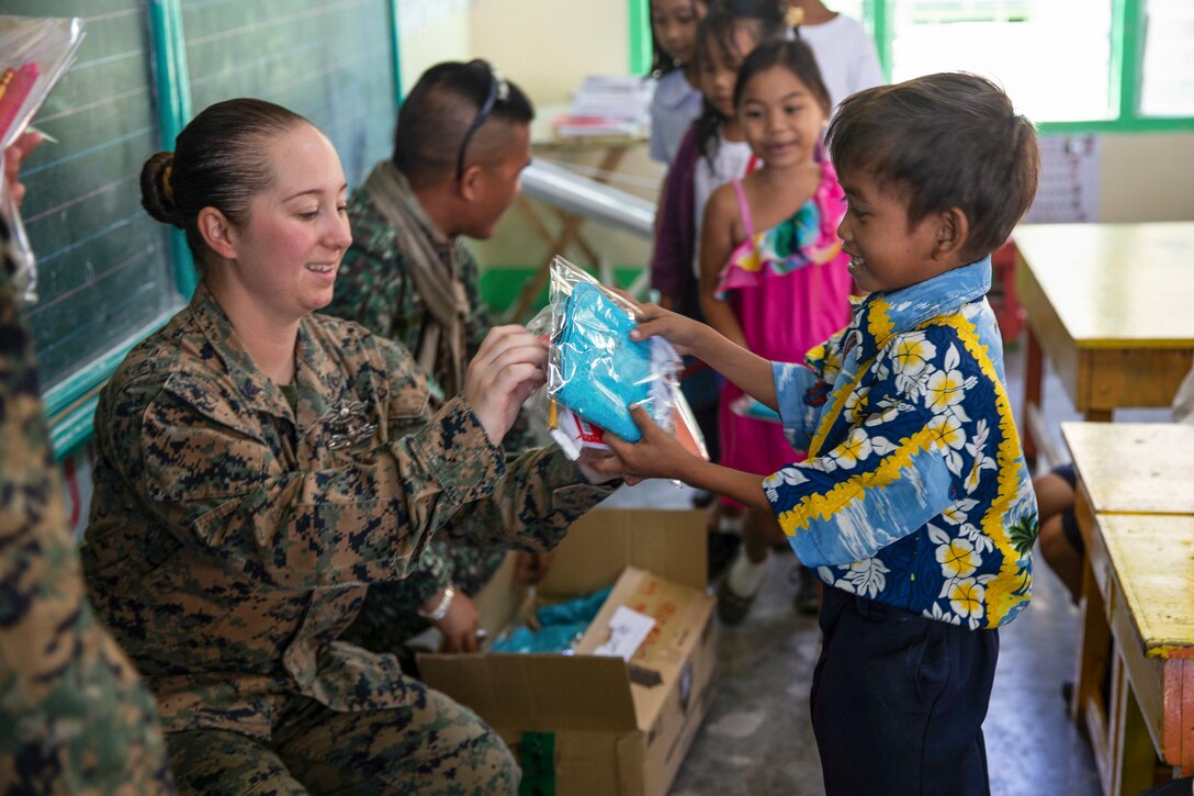 A sailor passes a gift to a smiling child, as other children and service members gather in the background.
