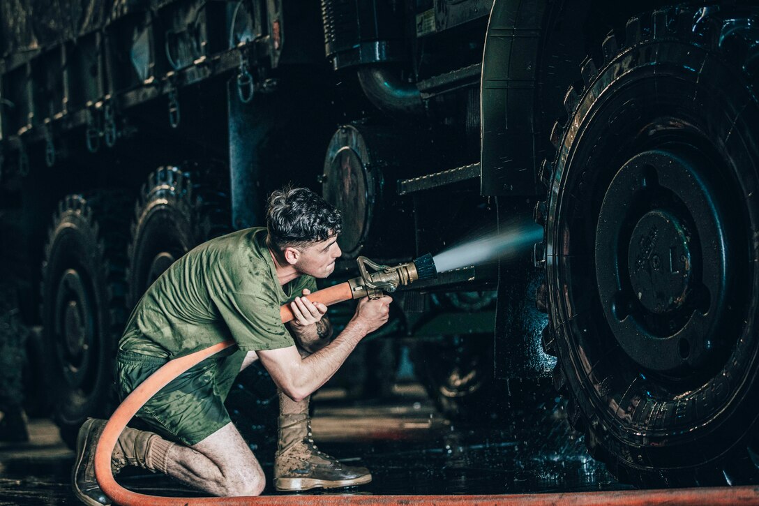 A kneeling Marine sprays water from a hose on a truck's underside.