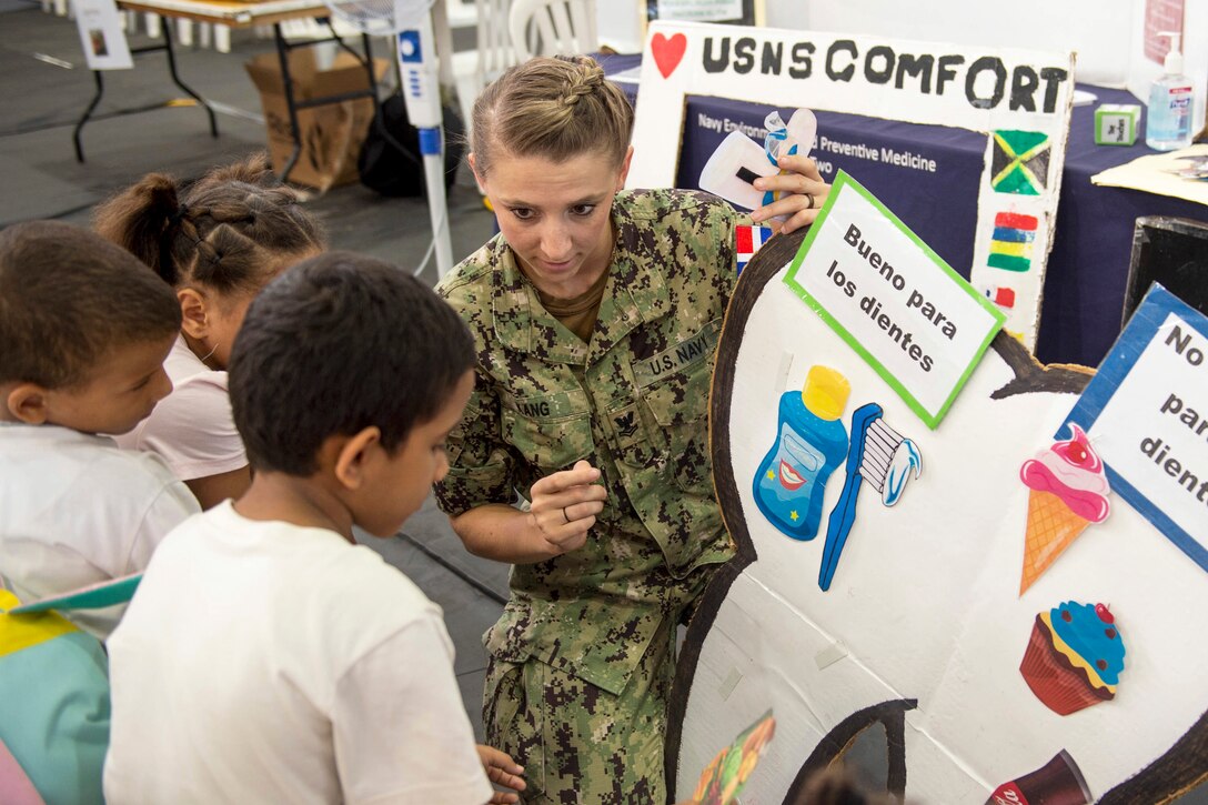 A sailor holds up a poster with illustrated information about dental health as children look on.