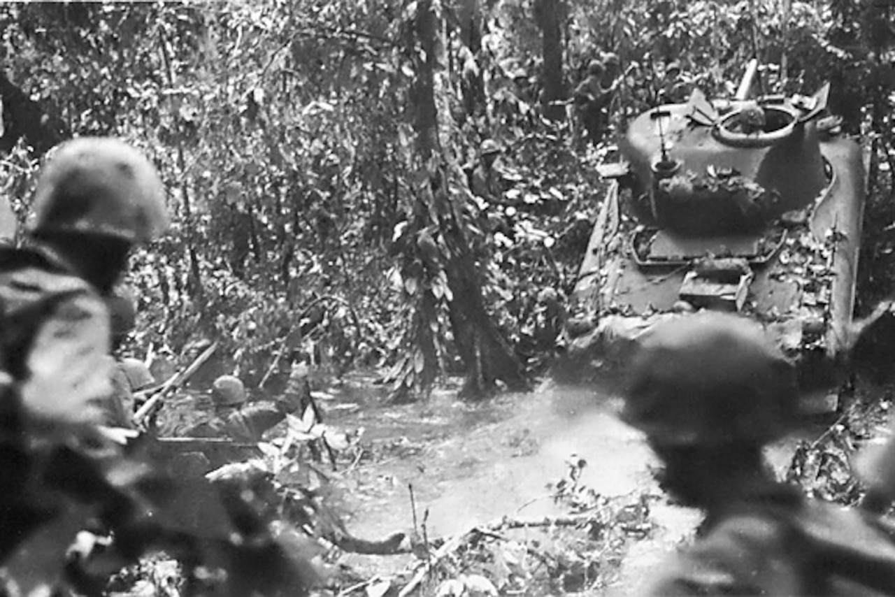 A tank emerges from a stream and up an embankment while several service members trail behind amid dense foliage.