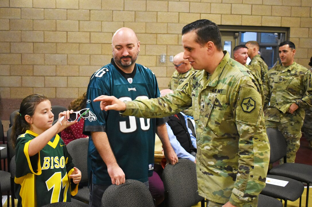 A military service member interacts with a young girl, who has a large pair of plastic sunglasses.