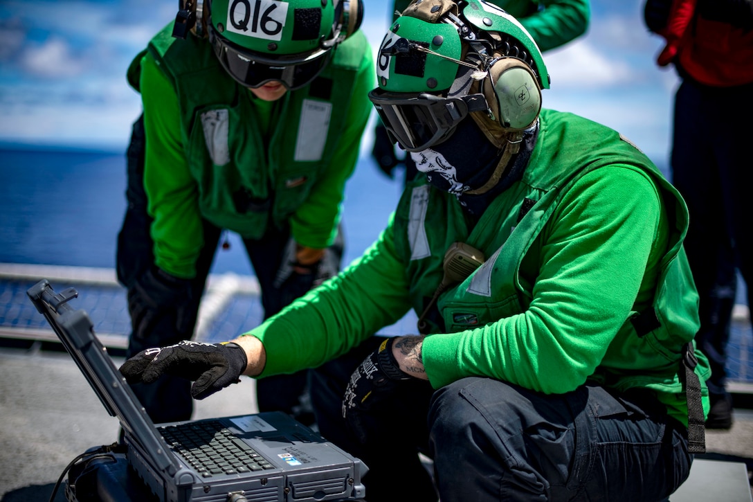 A sailor works on a laptop-type device on a flight deck as another watches.