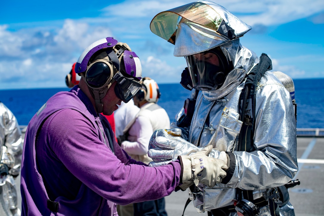 A sailor in purple helps another sailor put on gloves on a flight deck.