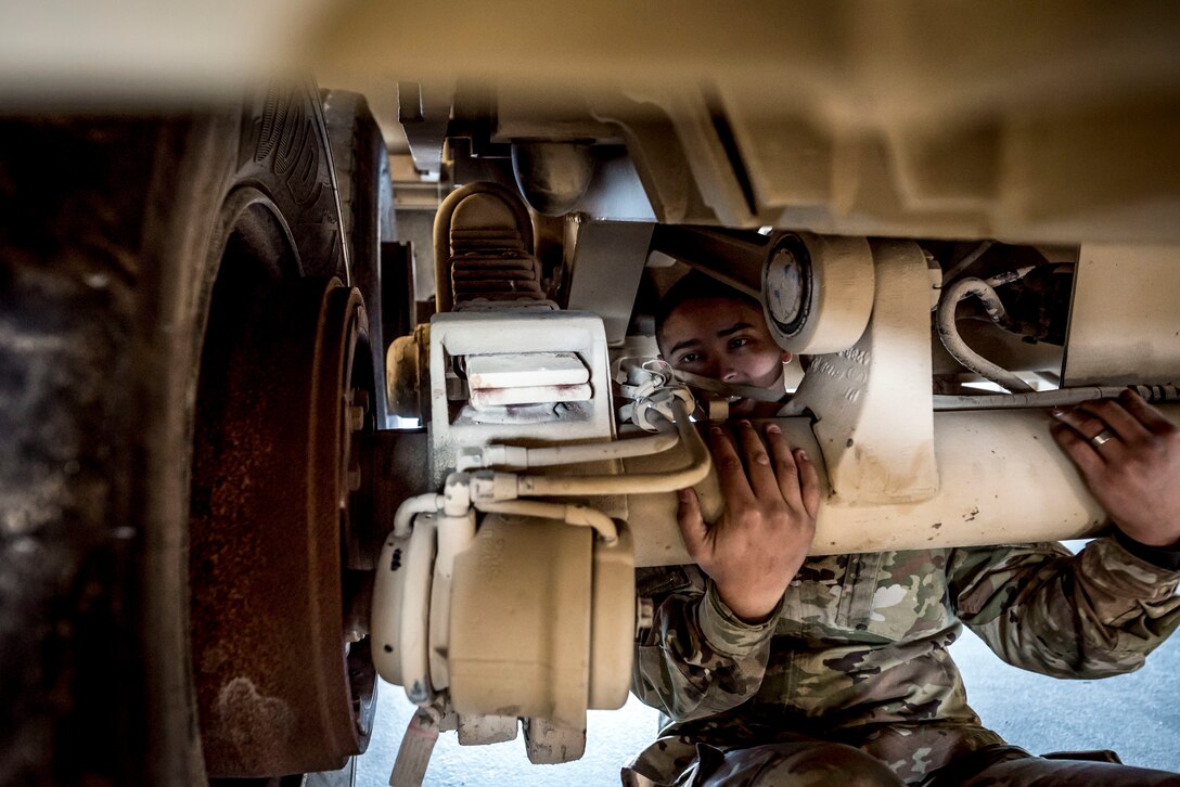 A soldier holds part of a vehicle's undercarriage while visually inspecting it.