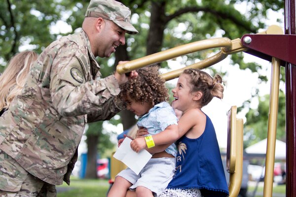 A service member interacts with his children who are seated on playground equipment.