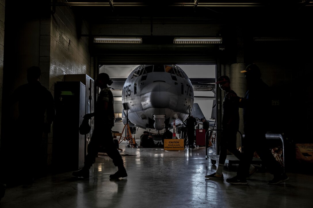 Four people walk across a hangar while others work on an aircraft in  the background.