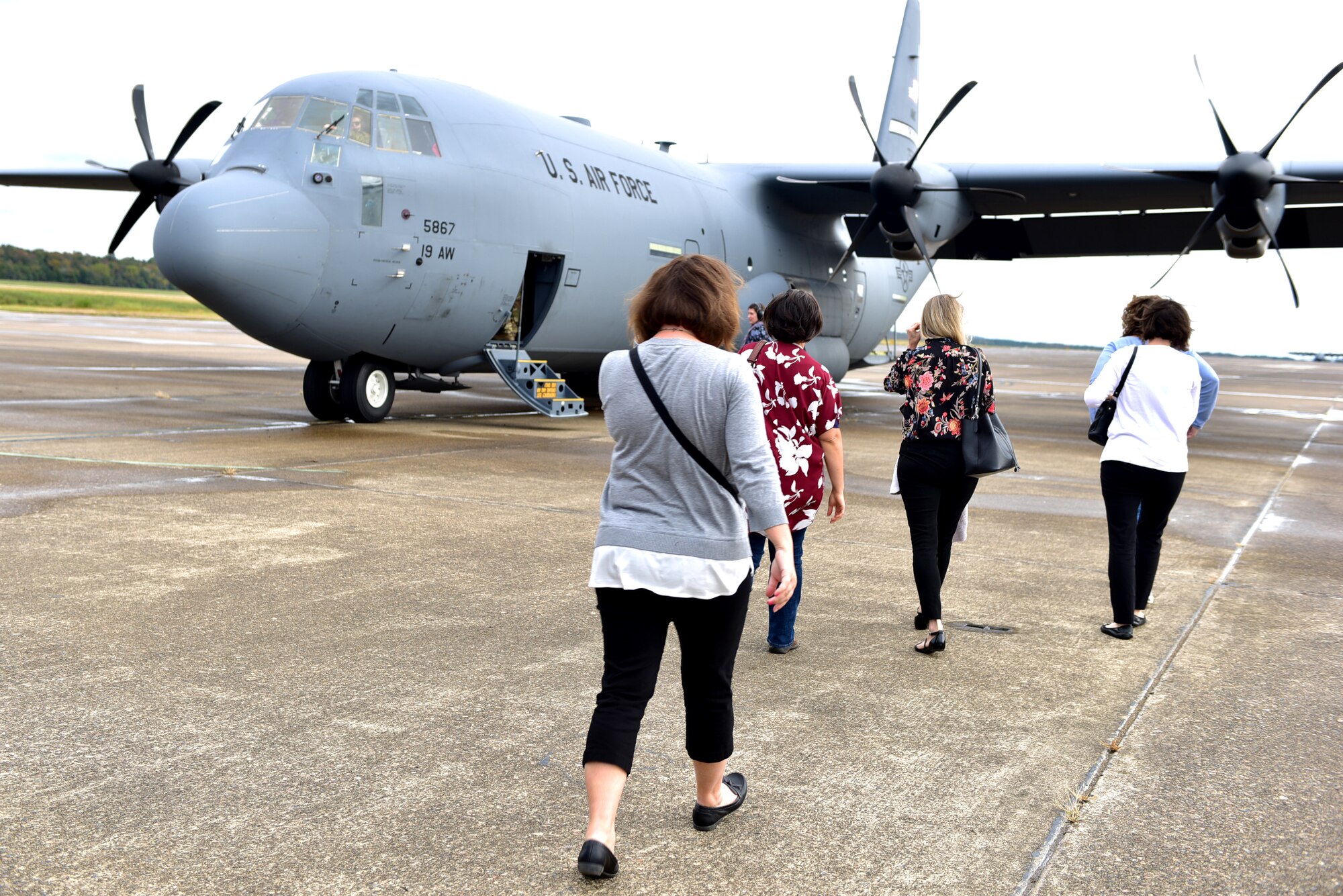 Females walk toward a large grey aircraft on a flight line. The sky is bright and the women are shielding their faces.