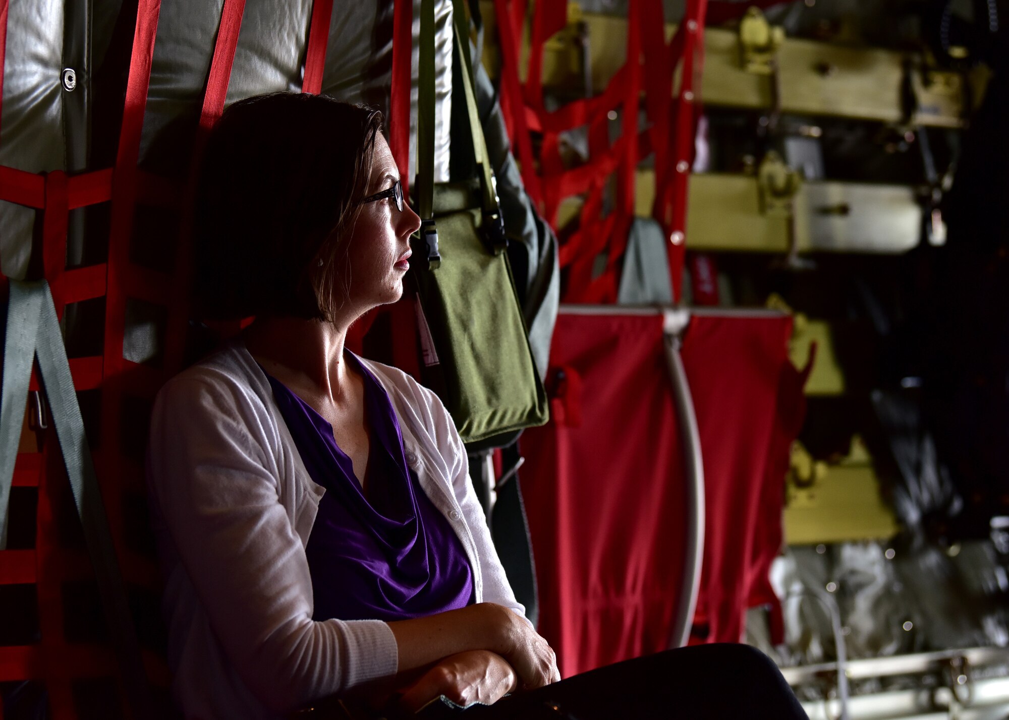 A female sits on red seats inside a grey aircraft. The womans face is lit up by the outside light and her hands are crossed in her lap.