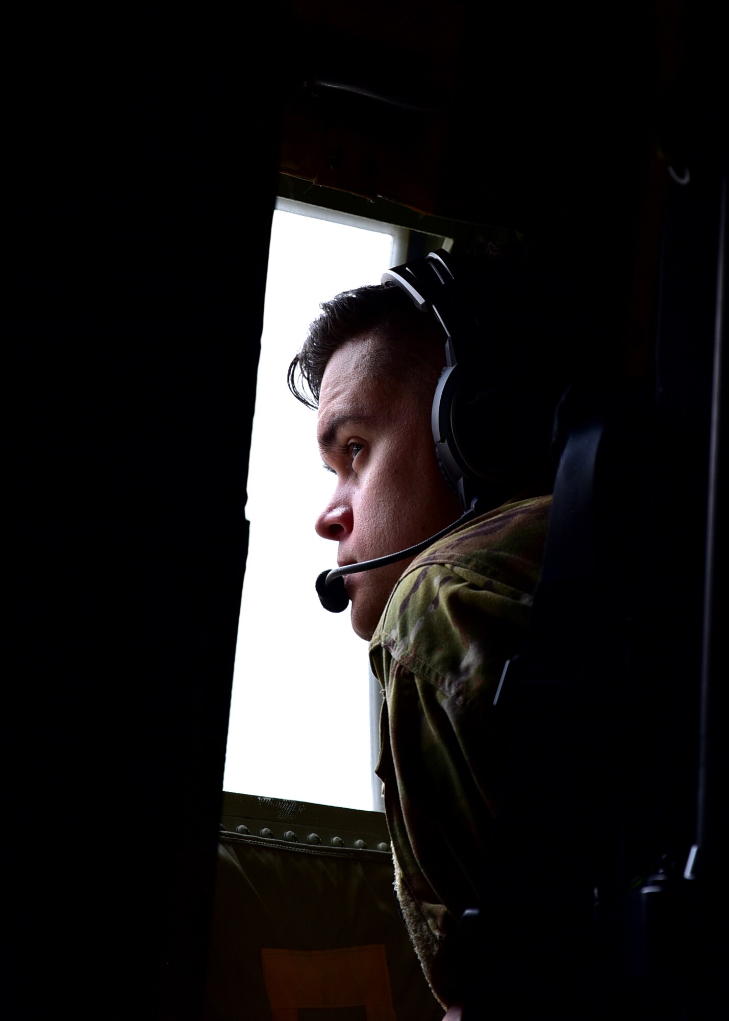 A male wearing a headset leans toward an airplane window in a dark cabin, his face is lit up by the outside light.