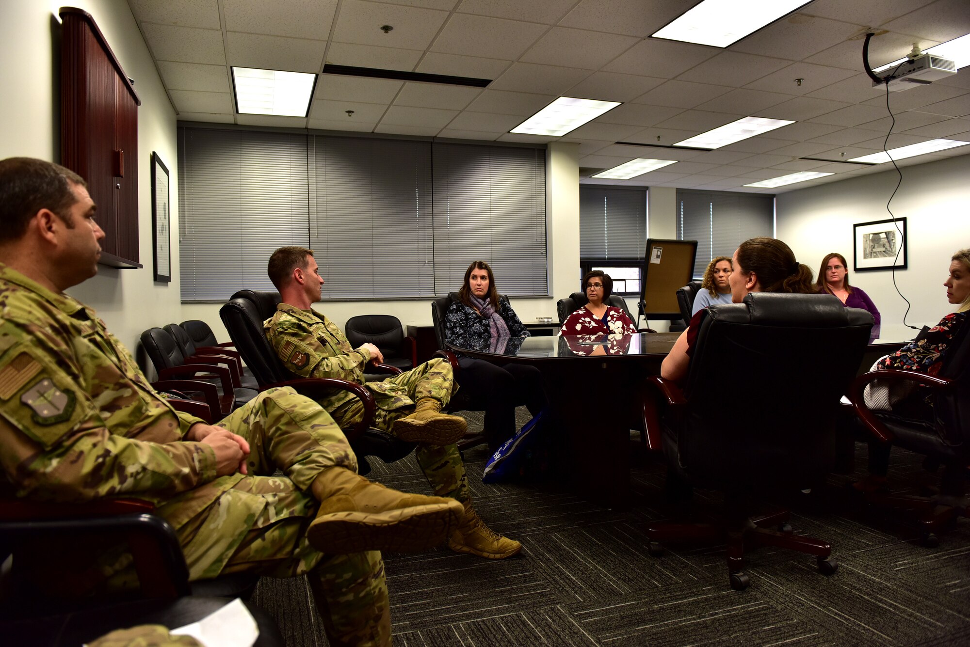 Individuals recieve a briefing around a round table in a biege room. A man in uniform is at the front of the table addressing people in civilian clothing.