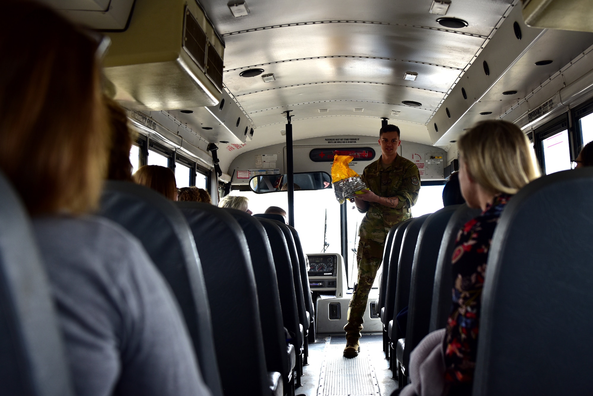 Individuals recieve a briefing inside a bus with rows of seats. The briefer is holding a yellow oxygen bag for demonstration purposes.