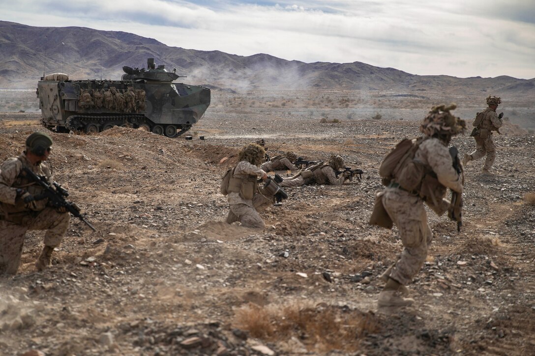 Several Marines carrying weapons move forward on desert-type terrain near an amphibious vehicle.
