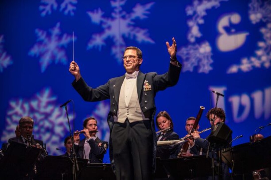 Lt. Col. Daniel Price turns to the audience during a holiday concert to conduct the audience in singing along with the Concert Band.  Academy Band musicians perform under a huge blue screen covered in snowflake designs behind him.