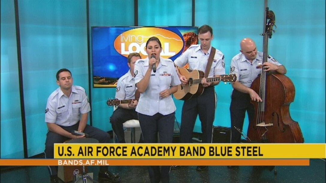 Five Air Force musicians perform in their short sleeved blue uniforms with guitars, upright bass, shaker percussion, and vocals on a "Living Local" segment for a television station.