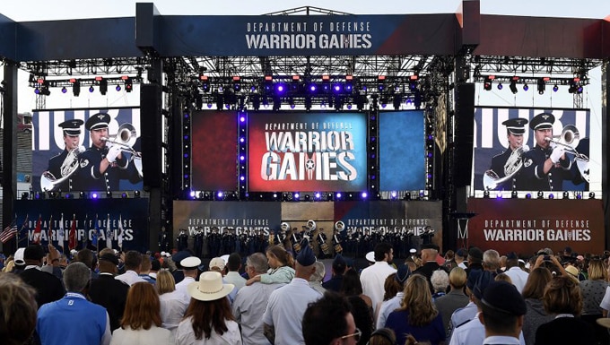 Musicians from the Academy Band marching band perform in blue uniforms on a large stage for a huge crowd for the opening ceremony of the Warrior Games.  Massive television screens on either side zoom in on trombone and horn players.