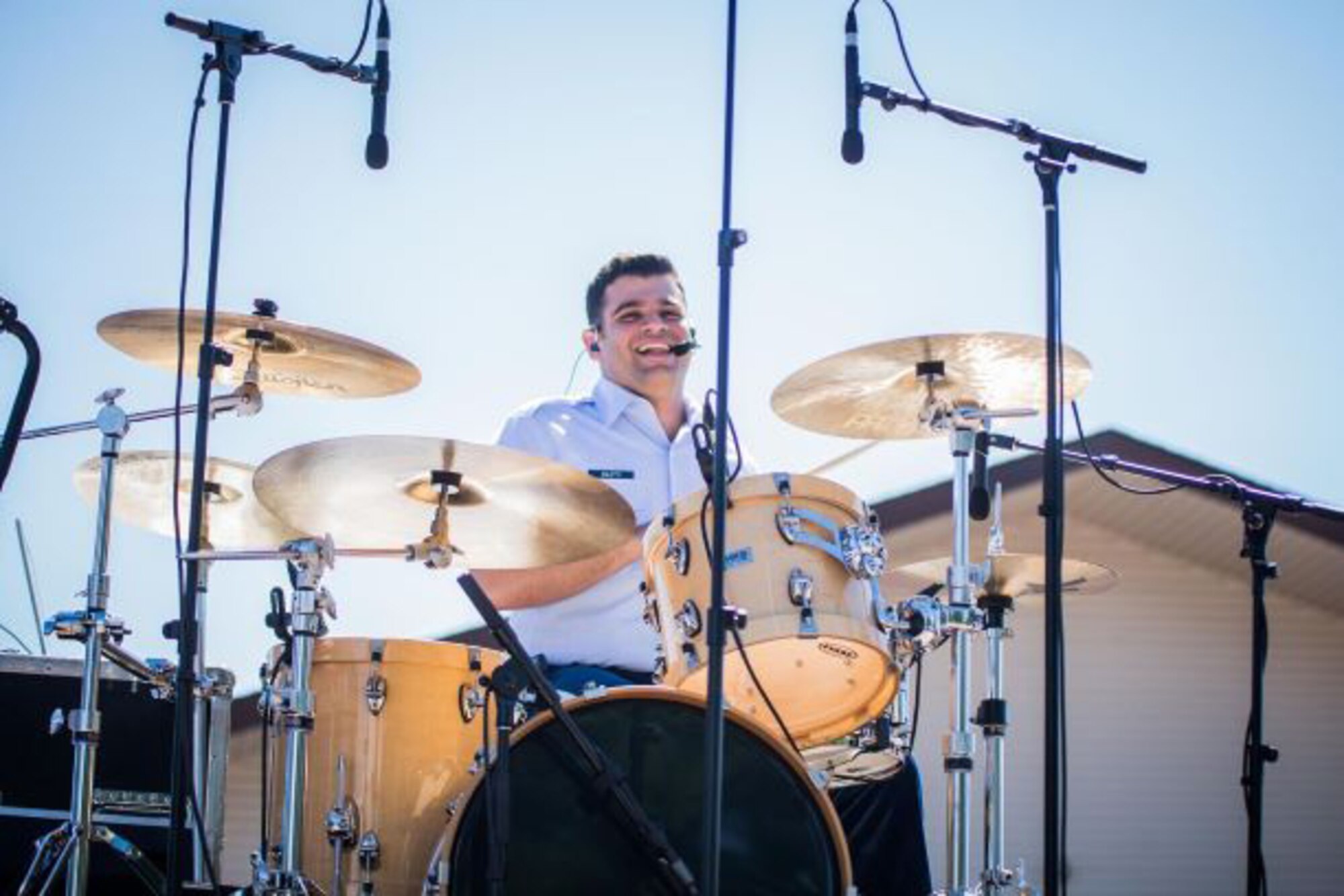 An Air Force musician joyfully performs on a drum set in a light blue uniform outdoors as part of a country band concert.