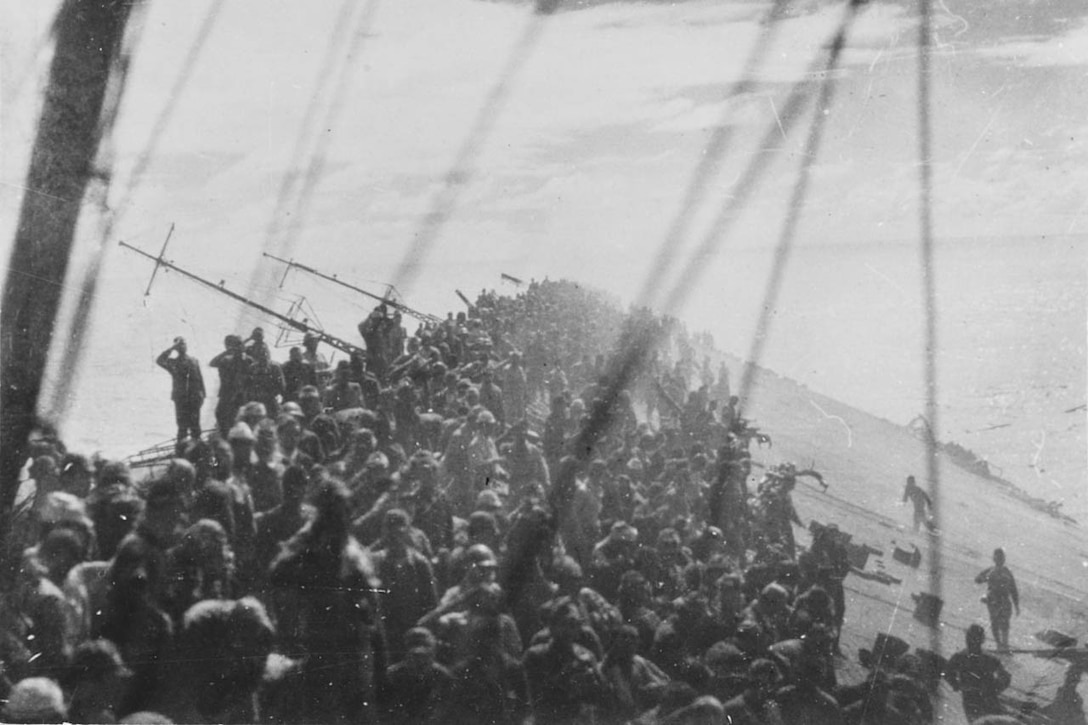 Men crowd the deck of a sinking aircraft carrier flight deck.