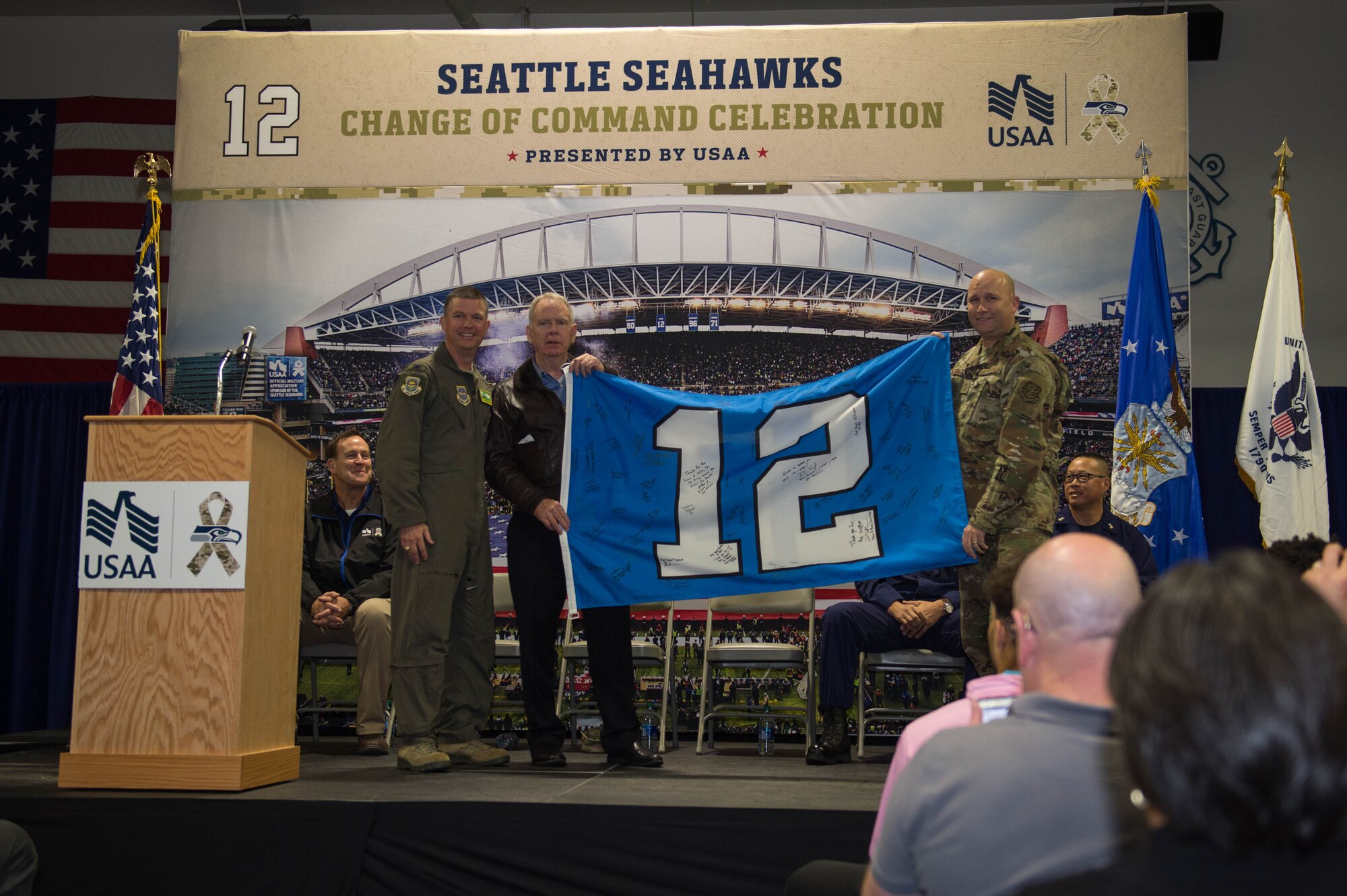 Col. Scovill Currin, 62nd Airlift Wing commander, left, Mike Flood, Seattle Seahawks vice president of community outreach, center, and Chief Master Sgt. Rob Schultz, 62nd AW command chief, right, display the 12th Man Flag during a “change of command” ceremony at Coast Guard Base Seattle, Wash., Oct. 22, 2019.
