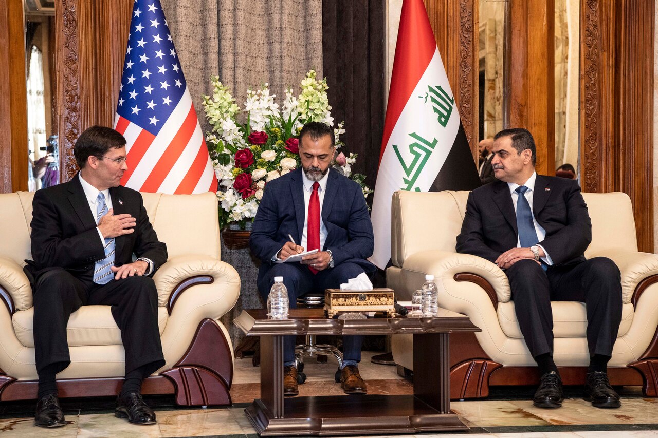 Defense Secretary Dr. Mark T. Esper sits in a chair and talks to a man seated next to him in an identical one, with a third man in between them taking notes.