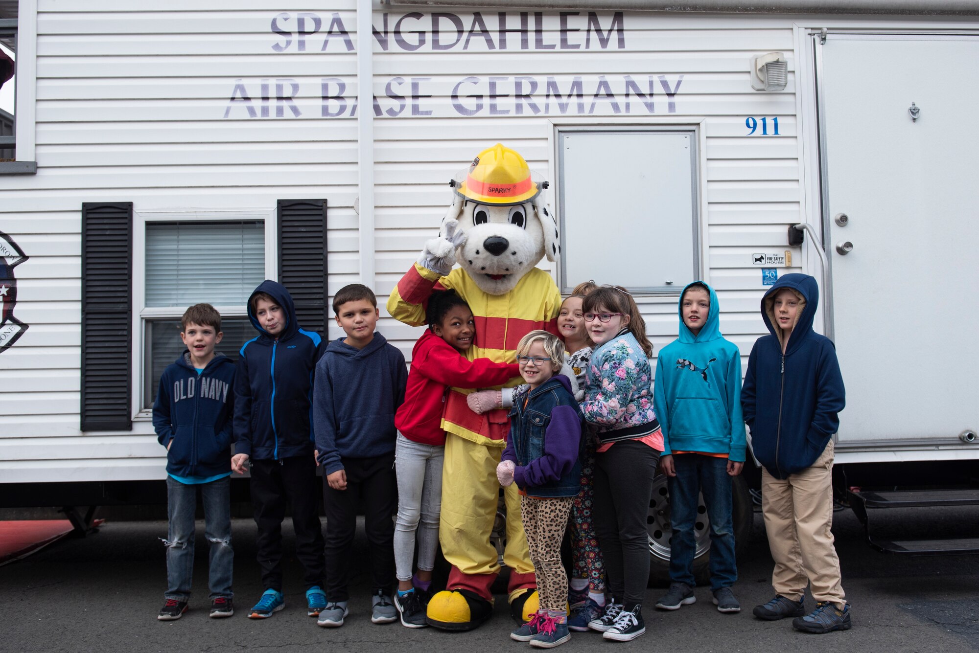 Students at Spangdahlem Elementary School pose for a photo with Sparky the Fire Dog outside the smoke demonstration trailer at Spangdahlem Air Base, Germany, Oct. 23, 2019. The smoke trailer provided students a first-hand experience on what to do in the case of a fire. (U.S. Air Force photo by Airman 1st Class Alison Stewart)