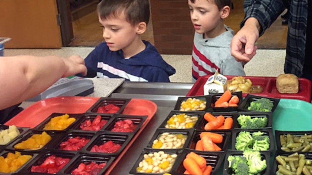Two students proceed through the lunch line with trays of small containers of a variety fresh fruits and vegetables