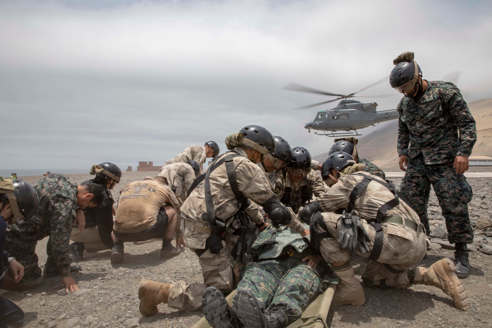Military personnel practice medical evacuations as a helicopter flies in the background.