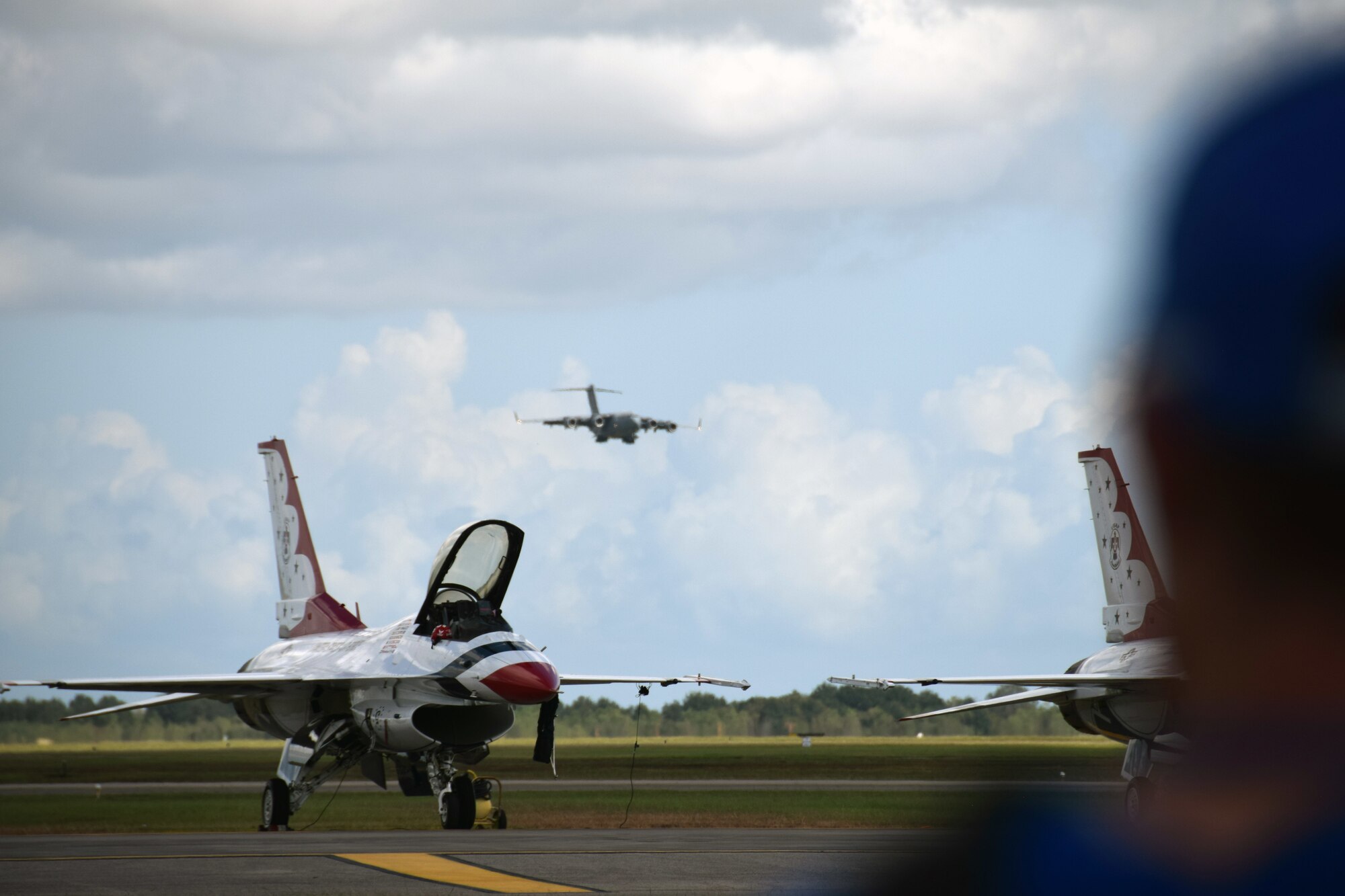 U.S. Air Force Thunderbird F-16C Fighting Falcons sit on the taxiway while a C-17 Globemaster III lands in the background at the Wings Over Houston Airshow at Ellington Airport, Houston Oct. 20, 2019.