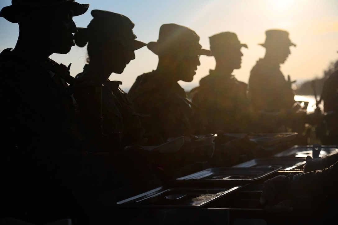 A silhouette of Marines standing in a line in front of a table with food on top.