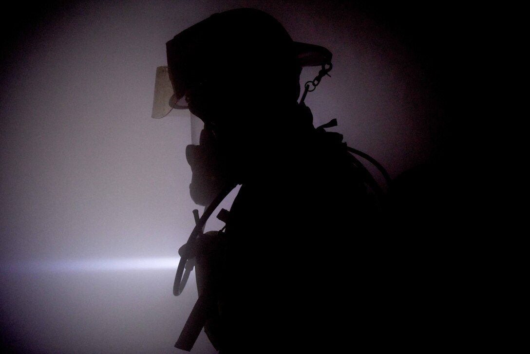 An airman wearing a firefighter uniform stands in a smoky room.