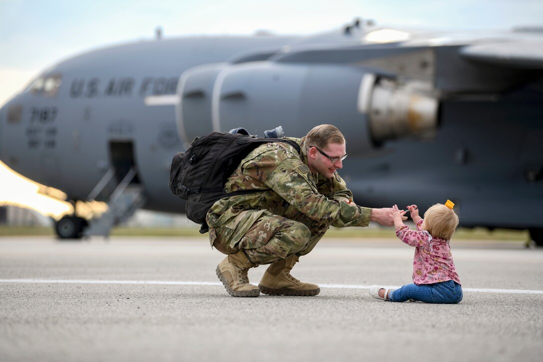 An airman kneels and reaches out his arm to interact with a baby, who is sitting on a flightline with a plane in the background.
