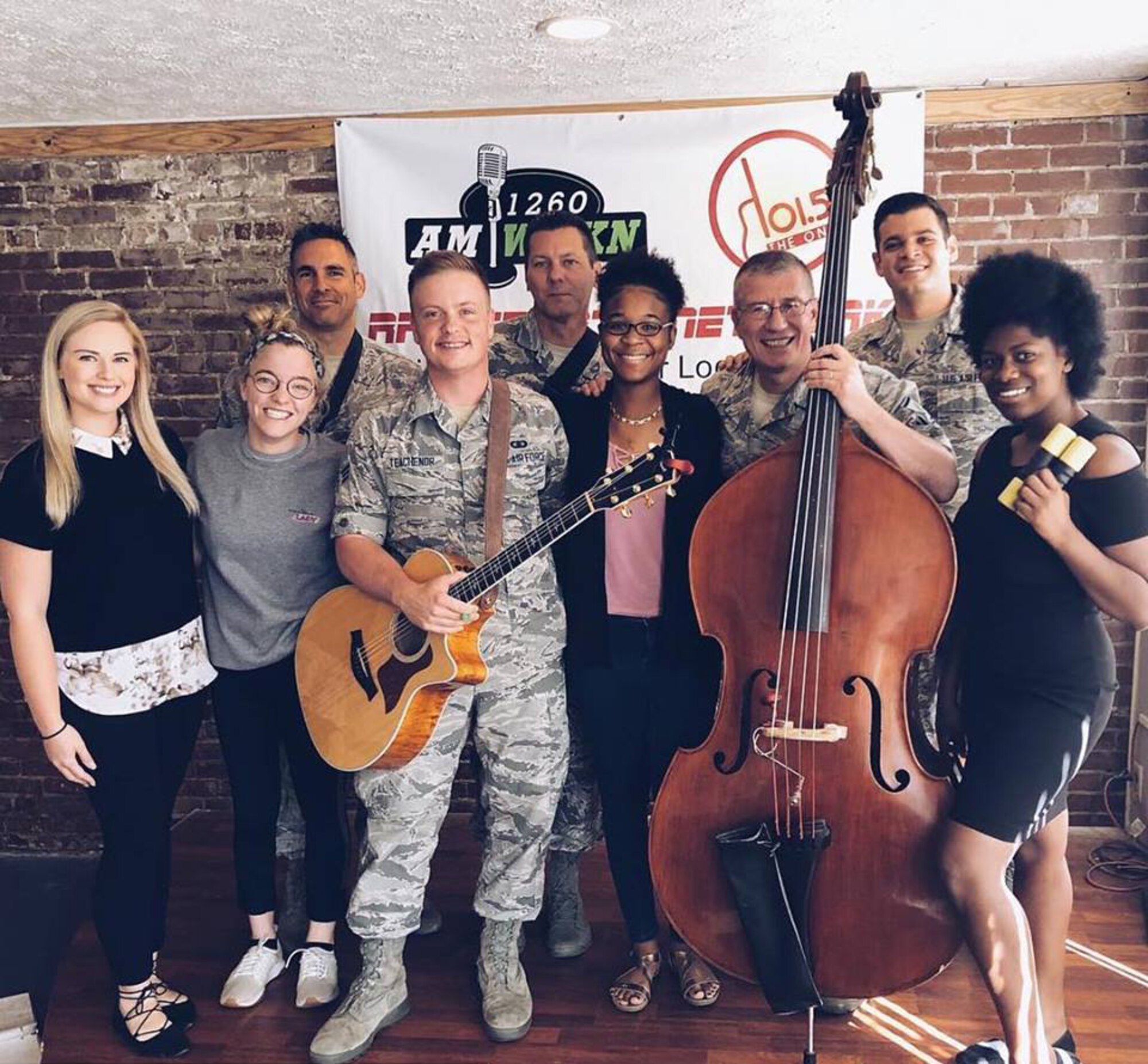 Five musicians from Wild Blue Country pose with their instruments, including an upright bass and electric guitar, in their digital camoflage pattern uniforms with local musicians and engineers.