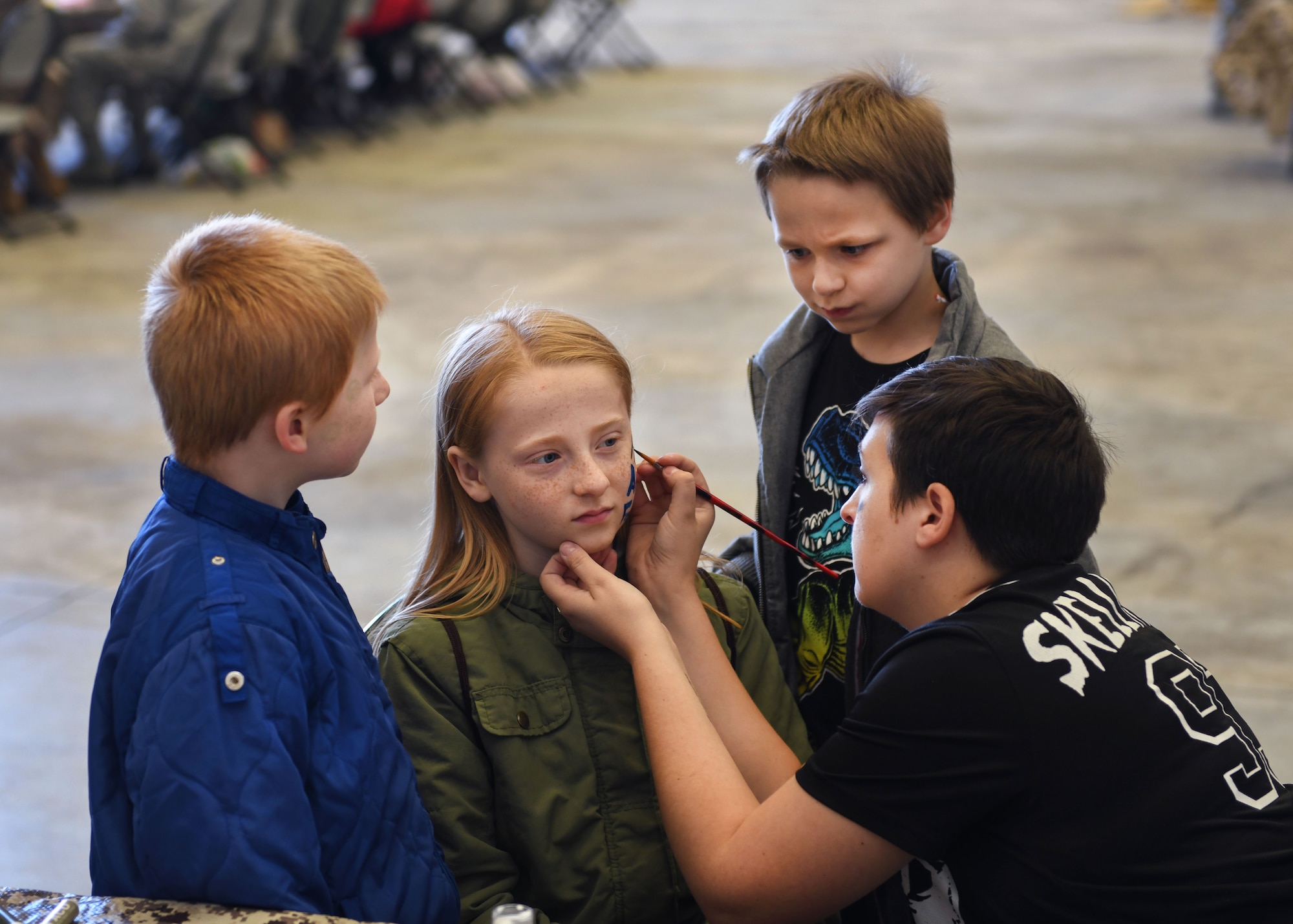 Two children watch as a girl gets her face painted by a volunteer.