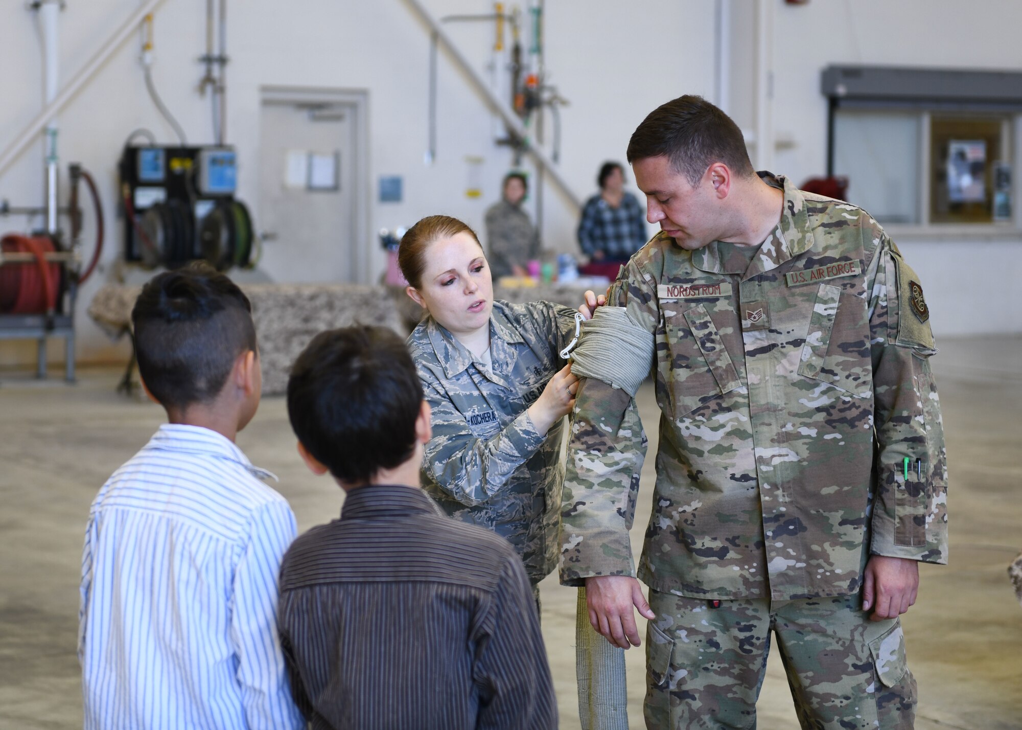 Two kids watch as an Airman demonstrates putting on a bandage on another Airman's arm.