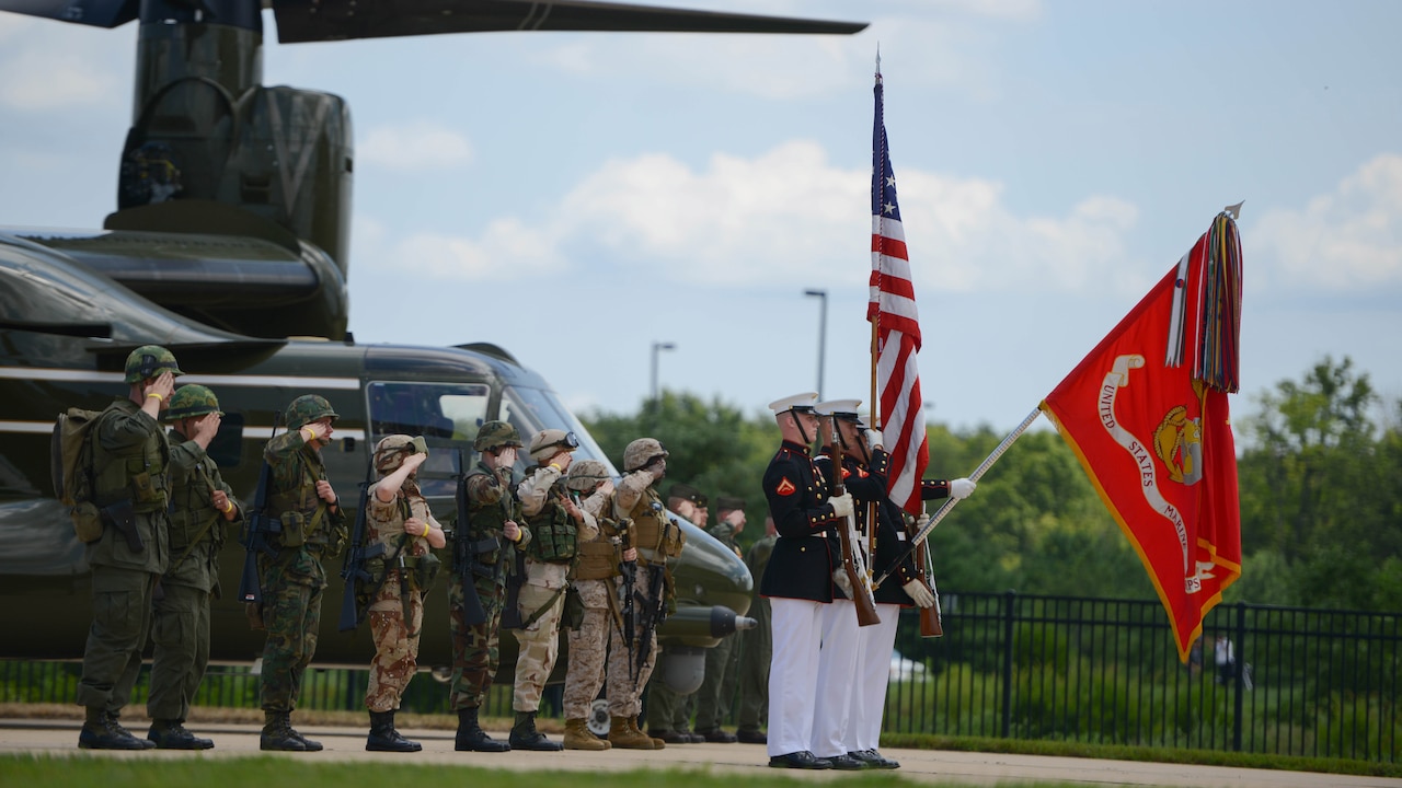 Marines in green and khaki uniforms and helmets salute in front of a helicopter and stand behind a Marine color guard with rifles, an American flag and a Marine Corps flag lowered.