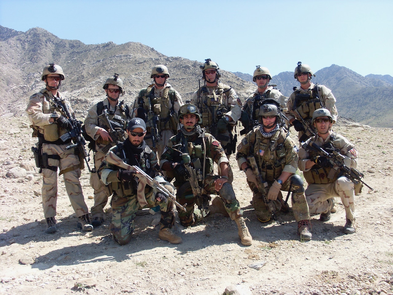 A group of soldiers dressed in full battle uniform pose together on the top of a mountain.