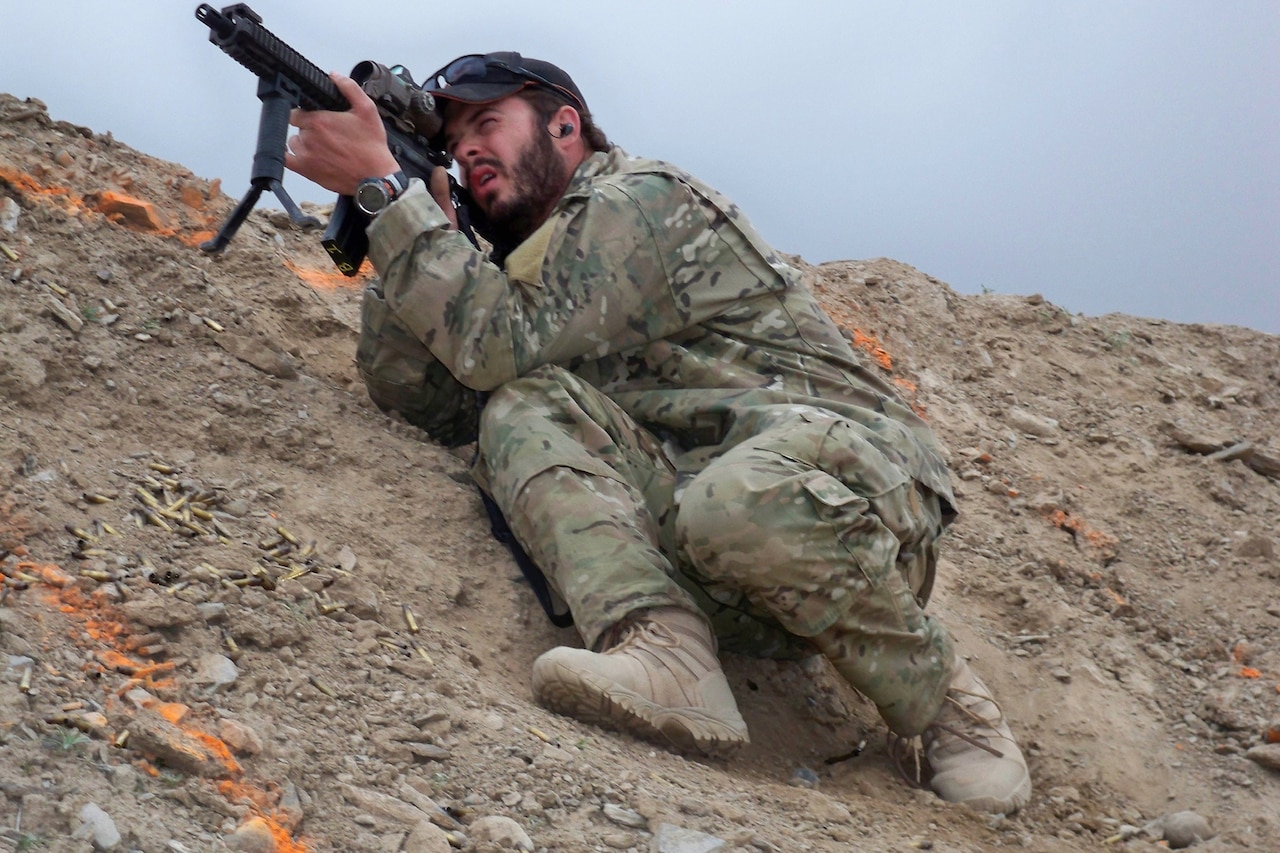 A soldier crouches on the side of a steep dirt mountain looking through the scope of his rifle