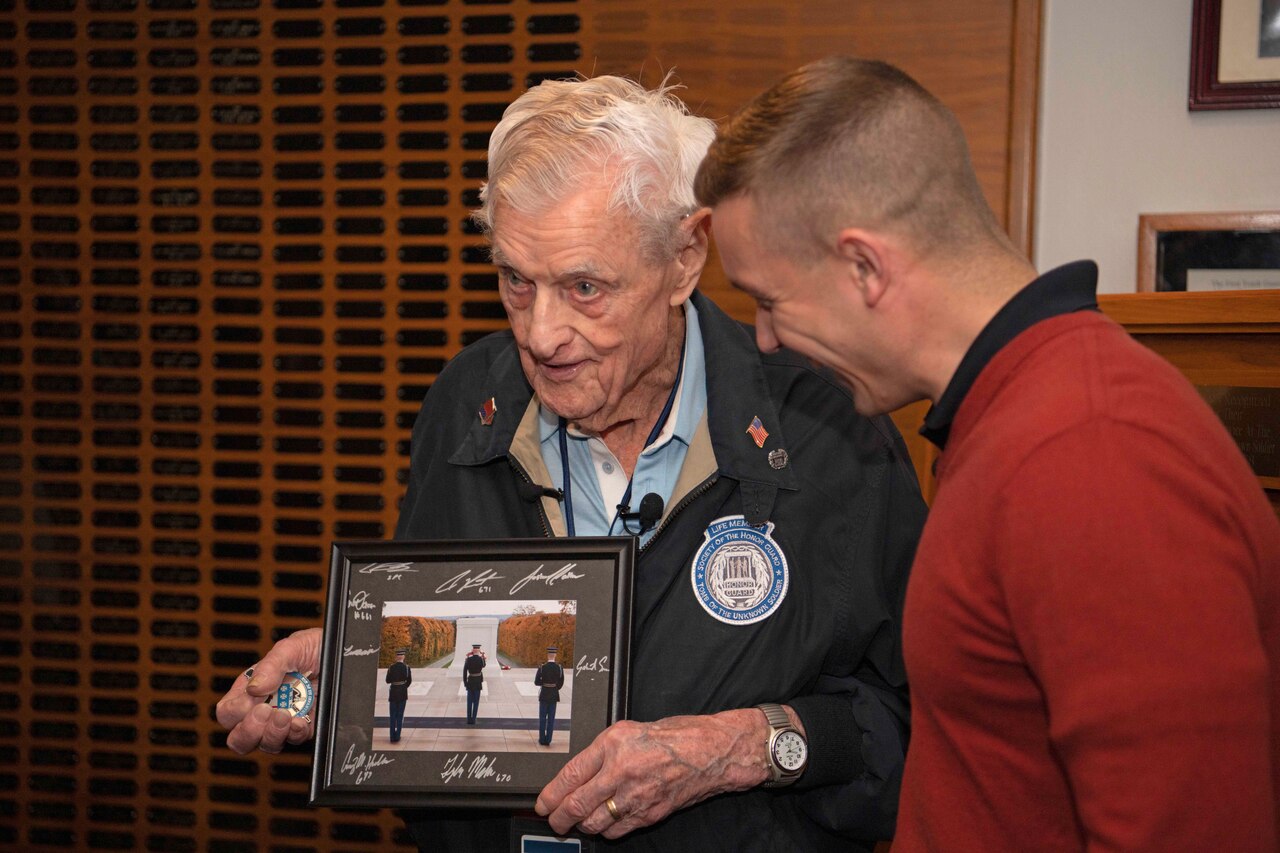 An elderly man holds a framed photo and a challenge coin as a younger man looks on.
