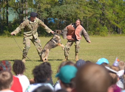Staff Sgt. Jenning Casey, training supervisor assigned to the 628th Security Forces Squadron, Staff Sgt. Timothy Merrigan, a military working dog handler assigned to the 628th SFS, and military working dog Szultan simulate an attack, October 23, 2019, at Marrington Elementary School on Joint Base Charleston, S.C. They were at the school to educate the children about how the military working dogs are trained to stop criminals and detect drugs. Security Forces personnel were at the school to educate students about drug prevention during Red Ribbon Week.  Red Ribbon Week, which takes place October 23-31, is an awareness program that began in 1985 as a way to show opposition to drugs as stated by the Get Smart About Drugs foundation.