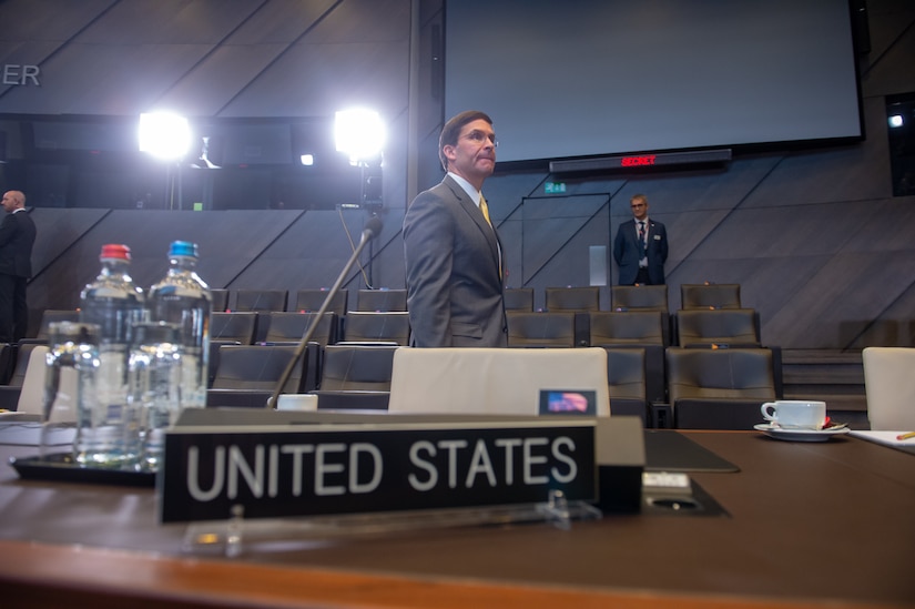 Mark Esper walks in front of bright camera lights, a United States desktop sign in the foreground.