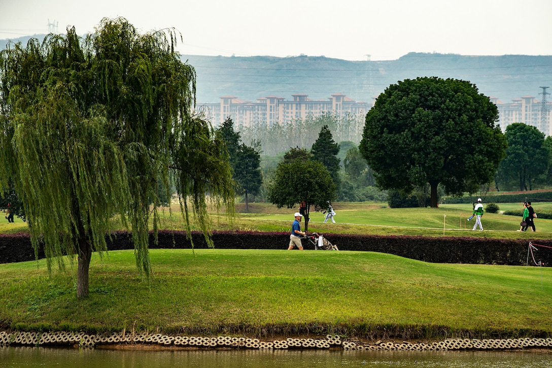 A golfer wheels his clubs next to a green, surrounded by the course’s scenic beauty, with trees on the course and mountains in the distance.