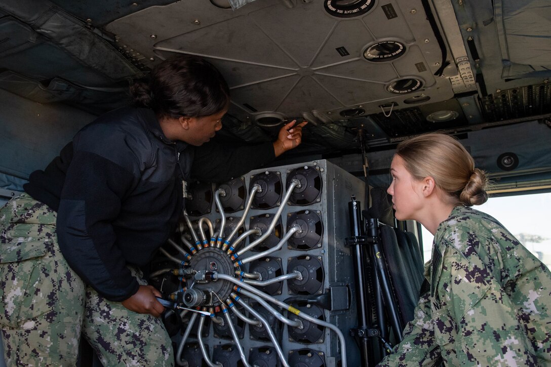 Two female service members work on the underside of a military helicopter.