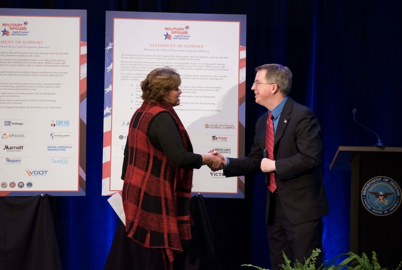 A man in a business suit shakes hands with a woman in a red plaid vest.