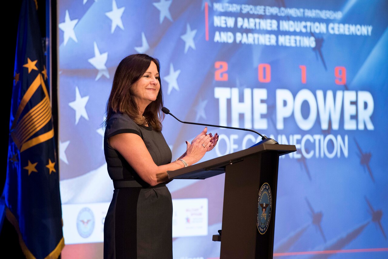 A woman standing behind a lectern applauds.