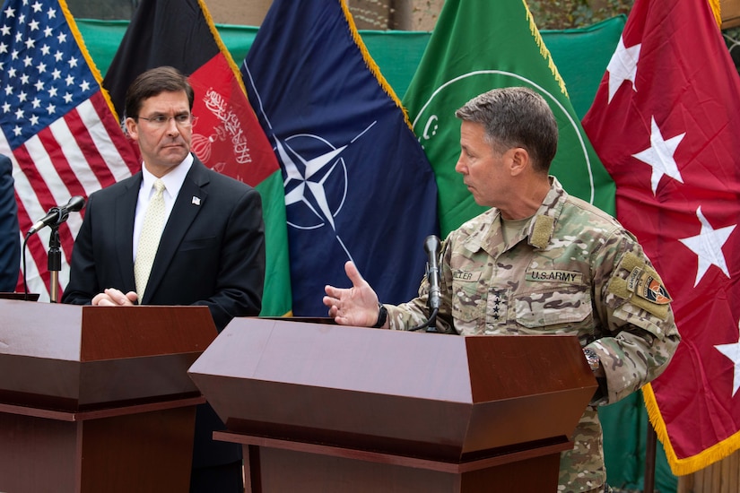 A man in civilian clothing and a man an Army uniform stand behind podiums with flags in the background.